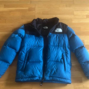 Blå dunjacka från The North Face - Säljer en blå dunjacka från The North Face med svart axelparti och logga på bröstet. Jackan har dragkedja framtill, två sidofickor och en hög krage. Perfekt för kalla dagar och har en cool, puffig look som är riktigt trendig. Säljer då den tyvärr är för liten.. 