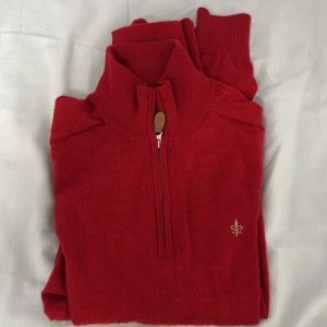 Röd merinoullströja från Morris XS - Riktigt snygg röd stickad tröja från Morris i mjuk merinoull. Halv dragkedja med läderdetalj och broderad logga på bröstet. Hög krage och långa ärmar, perfekt för kyliga dagar. Klassisk och stilren modell som är lätt att matcha.