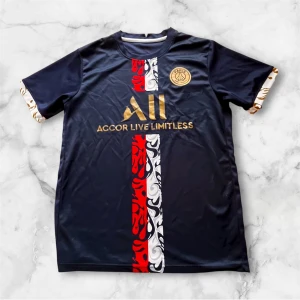 PSG fotbollströja marinblå guld - Snygg PSG fotbollströja i marinblått med guldiga detaljer och klubbmärke på bröstet. Framsidan har ett vertikalt mönster i rött, vitt och svart med grafiska former. Kortärmad modell med rund hals och tryckta detaljer på ärmsluten. Materialet är lätt och andas, perfekt för match eller träning.