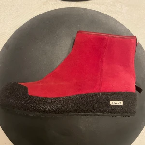 Röda mockaboots från Bally EU40 - Säljer ett par röda boots från Bally i mocka med grov svart gummisula och rund tå. Insidan är fodrad med mjukt vitt material för extra värme. Snyggt Bally-märke på sidan och minimalistisk design. Perfekt för dig som gillar statement-skor med lyxig känsla.