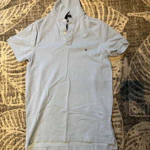 Ljusblå polo t-shirt Tommy Hilfiger  - Säljer en ljusblå polo t-shirt från Tommy Hilfiger i slim fit-modell. T-shirten har korta ärmar, klassisk krage med knappar och den ikoniska Hilfiger-loggan broderad på bröstet. Tillverkad i mjuk bomull för en skön känsla. Går att förhandla om priset!