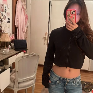 Svart croppad zipjacka - Svart croppad jacka med dragkedja framtill och lång ärm. Snygg, slim passform och tillverkad i ett mjukt, syntetiskt material som känns skönt mot huden. Perfekt att slänga på sig över en t-shirt eller linne. Enkel design utan mönster eller detaljer.