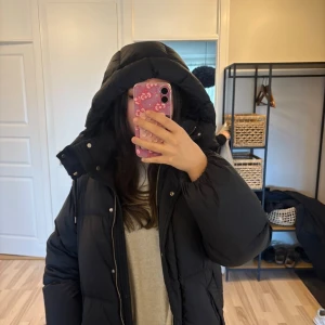 Svart pufferjacka från GANT - Svart pufferjacka från GANT med stor huva och snygga detaljer som logga på ärmen. Jackan har dragkedja framtill och är riktigt fluffig och varm, perfekt för kalla dagar. Passformen är normal och materialet känns slitstarkt och mjukt. (Priset kan diskuteras) ordinarie pris är 5000 sek.