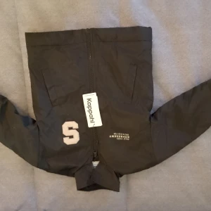 Svart vindjacka från Kappahl med tryck - Svart vindjacka från Kappahl med coolt S-tryck på bröstet och texten 'Be Your Best' med handgrafik på ryggen. Jackan har dragkedja framtill, ståkrage och två sidofickor. Perfekt för dig som gillar streetstyle och vill sticka ut med snygga detaljer.