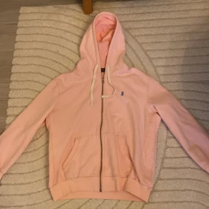 Rosa hoodie från Polo Ralph Lauren - Snygg ljusrosa hoodie från Polo Ralph Lauren med blå logga på bröstet. Tröjan har huva med snörning, dragkedja framtill och två fickor. Tillverkad i mjuk bomull, perfekt för chill dagar. Klassisk och stilren design som alltid funkar.