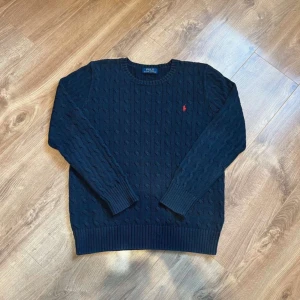 Mörkblå kabelstickad tröja Polo Ralph Lauren - Kabelstickad mörkblå tröja från Polo Ralph Lauren med klassisk rund halsringning och röd broderad logga på bröstet. Tröjan har långa ärmar och ribbade muddar för en snygg passform. Perfekt för dig som gillar stilren och tidlös design.