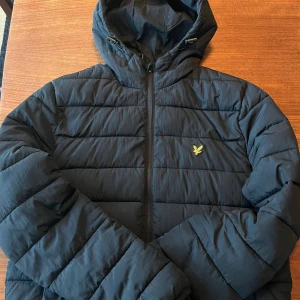 Svart pufferjacka från Lyle & Scott - Svart pufferjacka från Lyle & Scott med huva och gul logga på bröstet. Jackan har horisontella quiltade sektioner, dragkedja framtill och är långärmad. Perfekt för kyliga dagar och har en clean, modern look.
