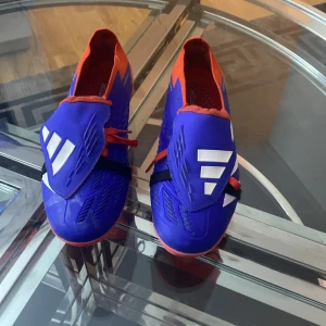 Adidas Predator blå/röda fotbollsskor - Säljer ett par Adidas Predator fotbollsskor i blått och rött med vita detaljer. Skorna har snörning, täckt plös och mönstrad sula med dobbar för gräsplan. Hybridtouch 2.0-material ger en skön känsla och bra grepp. Snygg design med Predator-logga under sulan.