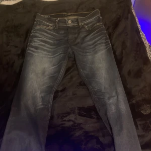 Mörkblå jeans från Jack and Jones  - Säljer ett par mörkblå jack and Jones  med klassisk femficksdesign och snygga slitningar framtill. Jeansen har raka ben och normal passform, med läderpatch bak i midjan. Perfekta för dig som gillar en tidlös och stilren look. Nypris 900