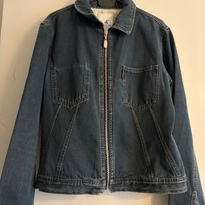 Mörkblå jeansjacka med dragkedja - Snygg mörkblå jeansjacka från Levi's med klassisk krage och två framfickor. Jackan har en rak passform och stängs med en grov dragkedja framtill. Perfekt för dig som gillar enkel och stilren denim med coola detaljer.