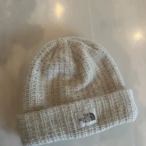 Beige stickad mössa från The North Face - Snygg beige mössa från The North Face med grovstickad utsida och mjukt fleecefoder på insidan. Klassisk uppvikt kant med logotyp framtill. Perfekt för kalla dagar och enkel att matcha med din vinterstil.
