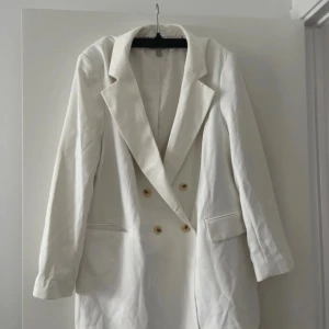 Vit kavaj med knappar - Snygg vit kavaj med ljusa knappar framtill samt på ärmsluten. Klassisk krage och två fickor framtill. Perfekt för dig som vill ha en stilren och fräsch look. Materialet känns slätt och lätt, troligen polyester eller bomull.