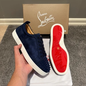Louboutin skor blå - Snygga marinblå sneakers från Christian Louboutin i mocka med vita sulor och den ikoniska röda undersidan. Skorna har snörning och coola nitar på tån för en edgy vibe. Perfekta för dig som vill sticka ut med lyxig streetstyle.