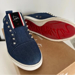Blå denim sneakers från Louboutin - Snygga blå sneakers från Christian Louboutin i denim med vita detaljer och ikonisk röd sula. Skorna har silverfärgade nitar längs snörningen och en vit perforerad insida. Perfekt för dig som vill sticka ut med exklusiv streetstyle. Väldigt fint skick och orders bekräftelse finns kvar. Dessa är omöjliga att få tag på i Sverige.