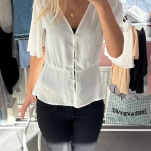 Vit blus från Gina Tricot - Säljer en vit blus från Gina Tricot med v-ringning och knappar framtill. Blusen har vida, korta ärmar och markerad midja för en feminin siluett. Tunt och lätt material som ger en luftig känsla.