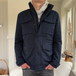 Marinblå fieldjacketfrån J.Lindeberg - Snygg marinblå fieldjacket från J.Lindeberg i storlek M. Jackan har flera praktiska fickor med lock, hög krage och dragkedja framtill. Materialet är slitstark polyester och passformen är normal. Perfekt för dig som gillar stilren och funktionell design.