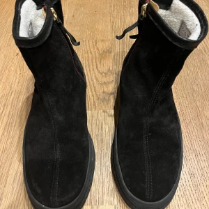 Svarta mockaboots i curlingmodell från Gant - Snygga svarta curling- boots från Gant i mjuk mocka med detaljer i skinn och dragkedja i guld på sidan. Fodrade med vitt foder för extra värme och komfort. Platt sula och stilren design som passar perfekt till höst och vinter.