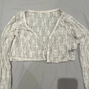 Vit spets bolerotopp - Säljer en vit bolerotopp i transparent spets med blommigt mönster. Toppen är croppad, har lång ärm och knappar framtill. Perfekt för att lägga till en extra touch till din outfit. Supersnygg att styla över linne eller klänning!