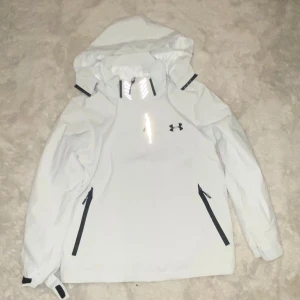 windbreaker  Under Armour - Snygg vit vindjacka från Under Armour med huva och svarta detaljer. Jackan har dragkedja framtill, två sidofickor med dragkedja och justerbara muddar med kardborre. Perfekt för blåsiga dagar och outdoor-aktiviteter.windbreaker 