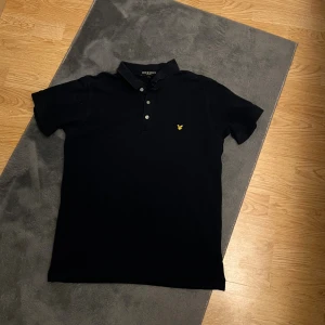 Svart pikétröja från Lyle & Scott - Klassisk svart pikétröja från Lyle & Scott med gul broderad logga på bröstet. Liten i storleken passar S bäst