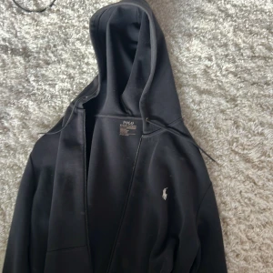 Svart hoodie från Polo Ralph Lauren - Svart hoodie från Polo Ralph Lauren med klassisk huva och dragkedja framtill. Tröjan har en vit broderad logga på bröstet och snörning vid huvan. Perfekt för chill dagar och enkel att matcha med jeans eller joggers.