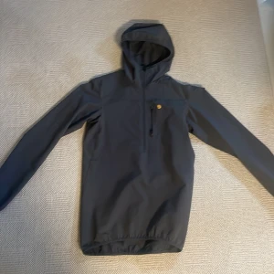 Svart vindjacka Fjällräven Bergtagen XS - Svart vindjacka från Fjällräven Bergtagen i herrmodell, storlek XS. Jackan har huva, lång ärm och en bröstficka med dragkedja. Materialet är slitstarkt och lätt, perfekt för uteliv. Klassisk gul Fjällräven-logo broderad på bröstet.