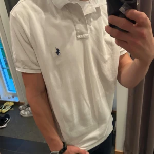 Vit pikétröja från Ralph Lauren - Klassisk vit pikétröja från Polo Ralph Lauren med blå broderad logga på bröstet. Tröjan har korta ärmar, krage och knappar framtill. Tillverkad i bomull med stretch för extra komfort. Perfekt för en clean och stilren look.