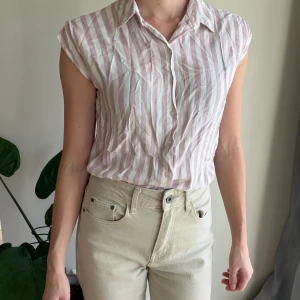 Randig ärmlös skjorta Vero Moda - Säljer en ärmlös skjorta från Vero Moda med breda vertikala ränder i ljusrosa och vitt. Skjortan har klassisk krage, knäppning framtill och är tillverkad i ett mjukt bomullstyg. 