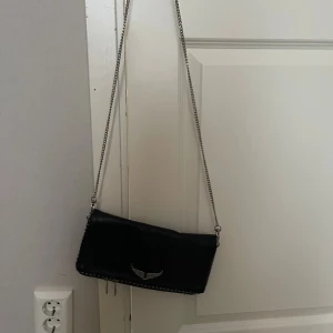 Zadig & Voltaire väska - Snygg svart axelväska i skinn med silverfärgad kedjerem och coolt vinge-emblem framtill. Väskan har ett fack  på baksidan och detaljer i silver. 