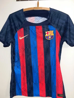 FC Barcelona matchtröja Nike - Säljer en officiell FC Barcelona matchtröja från Nike med klassiska vertikala blå och röda ränder. Tröjan har klubbmärket på bröstet och Nike-logga i guld. Tillverkad i lätt, ventilerande polyester med korta ärmar och rund hals. Perfekt för dig som älskar Barca!