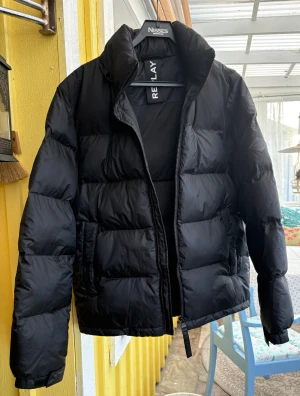 Svart pufferjacka från Replay - Säljer en svart pufferjacka från Replay med hög krage och dragkedja. Jackan är vadderad och har en clean look med Replay-logga på ryggen och armen. Perfekt för kalla dagar och har en riktigt skön passform. Materialet är syntet och jackan är designad för att hålla värmen. Mått finns på bild. Sonen som bär den på bilden är 187 cm lång. 