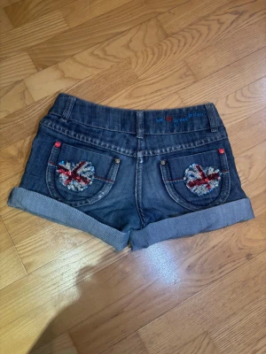 Blå jeansshorts med paljettdetaljer - Säljer ett par mörkblå jeansshorts med uppvikta ben och coola paljetthjärtan på bakfickorna i brittisk flaggdesign. Shortsen har röda och blå detaljer, broderad text 'we love great britain' och prickigt blått innerfoder. Perfekt för dig som gillar statement-plagg. kommer tyvärr inte till användning pågrund av storleken.💕