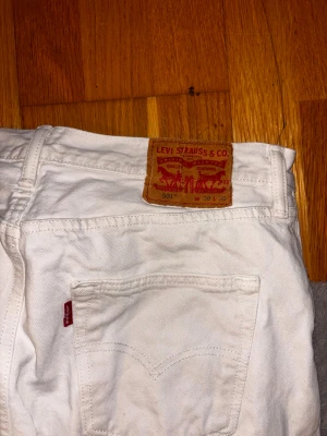 Vita Levi's 501 jeans straight fit - Säljer ett par vita Levi's 501 jeans med klassisk straight fit och knappgylf. De har fem fickor, Levi's röda etikett på bakfickan och brunt läderpatch i midjan. Perfekt för dig som vill ha en clean och tidlös look.