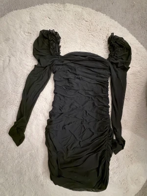 Svart långärmad puffärmklänning - Supersnygg svart kort klänning med långa puffärmar och rynkade detaljer längs hela kroppen. Klänningen har en offshoulder-design och volangdetaljer vid axlarna för en extra cool look. Perfekt för dig som vill sticka ut med en trendig och elegant stil.