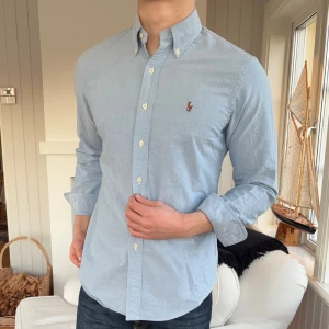 Blå skjorta från Ralph Lauren - Snygg ljusblå skjorta från Ralph Lauren med klassisk passform och button-down krage. Skjortan har långa ärmar, vita knappar och den ikoniska broderade loggan på bröstet. Tillverkad i mjuk bomull som känns skön mot huden. Perfekt för en clean och stilren look.
