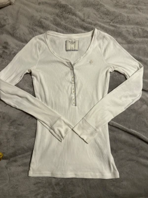 Vit långärmad Abercombie & Fitch topp med knappar - Supersöt abercombie and fitch henleytopp i storlek M. Bra men använt skick, vit. Har två små fläckar på ena ärmslutet (se bild) men inget som syns tydligt.❤️