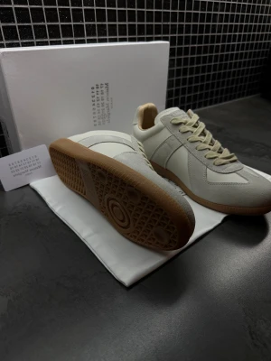 Maison Margiela gats - Maison Margiela sneakers i en stilren vit och beige färg med ljusbruna gummisulor. Skorna har rund tå, snörning och paneler i både skinn och mocka för en lyxig känsla. Perfekt för dig som gillar minimalistisk design och exklusiva detaljer.