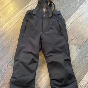 Svarta täckbyxor med hängslen - Svarta täckbyxor från Everest med justerbara hängslen, förstärkta knän och två dragkedjeförsedda fickor framtill. Byxorna har robust material och är perfekta för kalla dagar utomhus. Snygg och funktionell design med knappstängning i midjan.