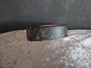 Svart Louis Vuitton bälte med LV-spänne - Snyggt svart bälte från Louis Vuitton med klassiskt LV-monogram och stort LV-spänne i metall. Bältet har en bred form och är tillverkat i präglat skinn med diskret mönster. Perfekt för att lyfta din outfit med en exklusiv touch.