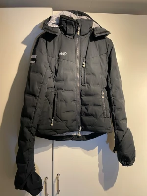 Marin ridjacka från Uhip  - RidJacka i figursydd och sportig modell med nedfällbara ärmslut som skyddar handen vid regn och blåst. Hög krage med fleecefoder. Två handfickor med fleecefoder och dragkedja samt en bröstficka med dragkedja. Dragsko i nederkant, luva och ärm. Luvan är dessutom hjälmanpassad. Jackan är som ny. Nypris på Hööks 1150:- 