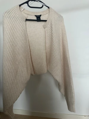 Beige stickad bolero från Lindex - En beige stickad bolero från Lindex i storlek M. Koftan har en öppen front och långärmade ribbade ärmar. Materialet är en mjuk mix av polyester, akryl och ull.  Obs: Alla plagg säljs endast fram till 28/12 