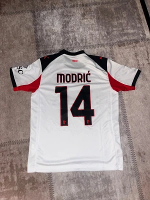 AC Milan Modric 14 vit matchtröja Puma - Säljer en AC Milan fotbollströja med Modric och nummer 14 på ryggen. Tröjan är vit med svarta och röda detaljer, svart krage och korta ärmar. Puma-logga på bröstet och ärmen, samt Emirates Fly Better tryck. Tillverkad i lätt polyester med DryCell och Re:Fibre teknologi.