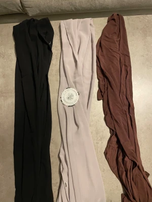 Tre enfärgade sjalar i olika toner - Säljer tre långa och tunna sjalar i svart, ljusbeige och brun. Alla är enfärgade och har en slät, mjuk yta som känns skön mot huden. Perfekt att styla runt halsen eller som hijab. Materialet är lätt och luftigt, troligen polyester eller bomull.