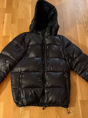 Svart pufferjacka från Polo Ralph Lauren - Svart pufferjacka från Polo Ralph Lauren med huva och flera dragkedjeförsedda fickor. Jackan har en röd Polo-logga på bröstet och quiltad design. Den är äkta!