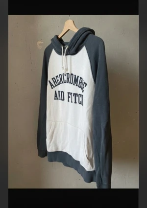 Abercrombie & Fitch hoodie i grått och vitt - Snygg hoodie från Abercrombie & Fitch med grå ärmar och huva samt vit framsida med stor logotyp. Tröjan har snörning vid huvan och en stor magficka. Perfekt för chill dagar och streetstyle. Mjuk bomullskänsla och långärmad modell.