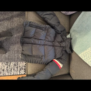 Moncler Chamonix S/M - Svart pufferjacka från Moncler med quiltad design och dragkedjor på fickorna. Jackan har en hög krage och insida i vit. På ena ärmen finns en röd och beige detalj med Moncler-logga. Perfekt för kalla dagar och riktigt snygg till streetwear.