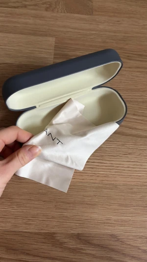 GANT glasögonfodral med putsduk - Snyggt och stilrent glasögonfodral från GANT i mörkgrått med matchande vit putsduk. Perfekt för att skydda dina glasögon och hålla dem rena. Fodralet har en mjuk insida och GANT-logga på både fodral och putsduk.