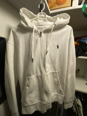 Vit hoodie från Polo Ralph Lauren - Vit hoodie från Polo Ralph Lauren med dragkedja, snörning i huvan och klassisk logga broderad på bröstet. Tröjan har två stora fickor framtill och är tillverkad i mjuk bomull. Perfekt för chill dagar och enkel att matcha med jeans eller joggers. har lite små hål som inte är så synliga 