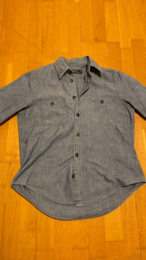 Blå skjorta från Polo Ralph Lauren - Snygg blå skjorta från Polo Ralph Lauren i storlek S. Skjortan har klassisk krage, två bröstfickor med knappar och knäppning hela vägen framtill. Tillverkad i mjuk bomull, perfekt för en chill och stilren look. Den är skön men samtidigt stil ren.