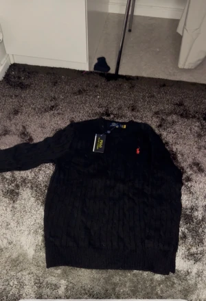 Svart stickad tröja från Polo Ralph Lauren - Svart stickad tröja från Polo Ralph Lauren med klassisk kabelstickad design och röd broderad logga på bröstet. Rund hals och långa ärmar, perfekt för dig som gillar stilrena och tidlösa plagg. Snygg ribbad kant vid ärmslut och nederkant.